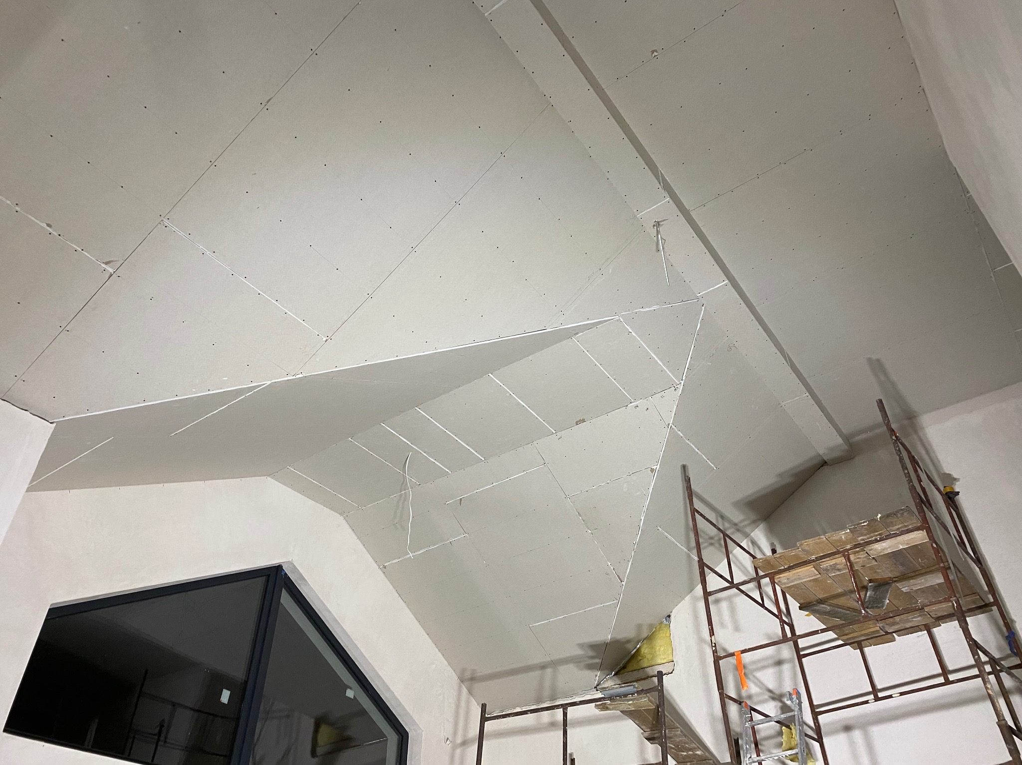 Skośny sufit z płyt karton-gips, widoczne łączenia i wkręty, rusztowanie w rogu. Ukończony etap prac w pomieszczeniu ze skośnym oknem.