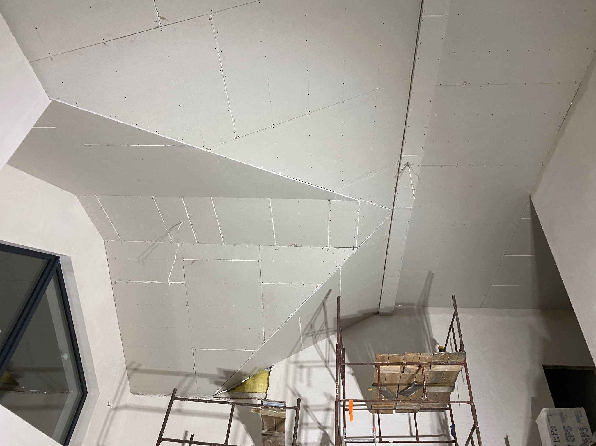 Skośny sufit poddasza z zamontowaną płytą kartonowo-gipsową, widoczne łączenia i wkręty, rusztowanie w pomieszczeniu, okno w tle. Prace wykończeniowe.