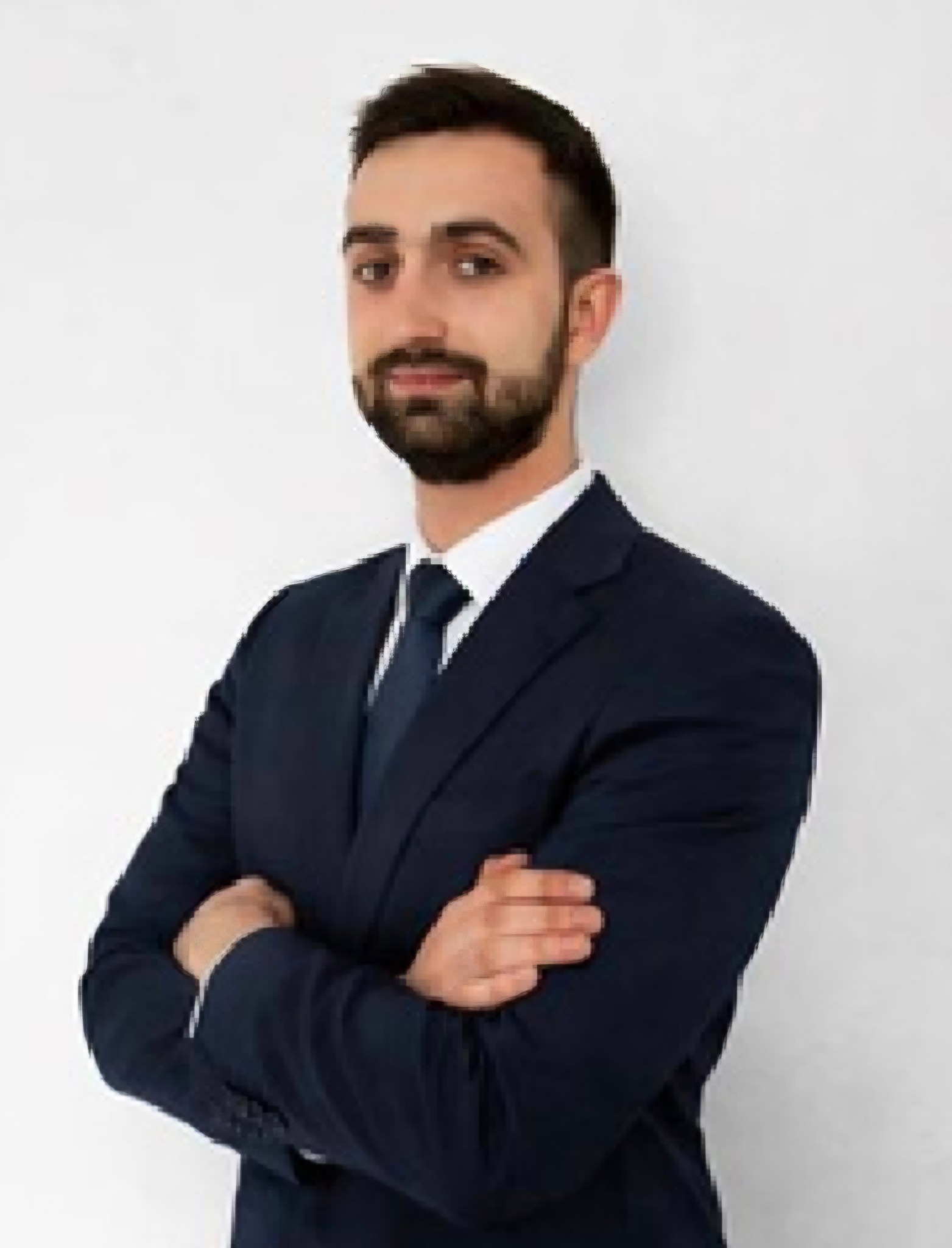 Młody mężczyzna z brodą, ubrany w granatowy garnitur i krawat, pozuje z założonymi rękami na tle białej ściany. Profesjonalny wizerunek.