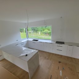 Kwarcowo - Nowoczesna kuchnia w trakcie wykańczania, z białymi szafkami i wyspą, wyposażona w jasne blaty z delikatnym użyleniem, częściowo pokryte folią ochronną. Duże okno z widokiem na zieleń.
