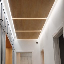 Meble na wymiar - Drewniany sufit z oświetleniem LED w korytarzu, widok z dołu. Po lewej szafa z szarymi frontami, po prawej białe ściany i drzwi. W tle widoczna biała ściana z płytkami.