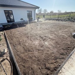 Aqua Garden - Przygotowany teren pod trawnik z widocznymi zraszaczami w trakcie nawadniania. W tle dom z ciemną stolarką okienną i zadbana zieleń.