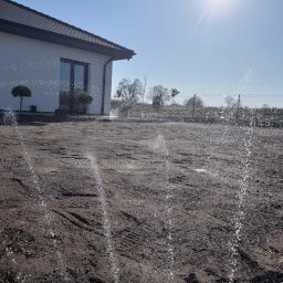 Aqua Garden - Automatyczne nawadnianie w nowo założonym ogrodzie. W tle biały dom z ciemnym dachem i drzewa. Woda tryska z dysz na przygotowaną glebę w słoneczny dzień.