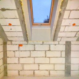 WJK Energia Wojciech Kubecki - Surowy stan poddasza z oknem dachowym i widocznymi przewodami elektrycznymi w ścianach z białego betonu komórkowego. Pomarańczowe puszki instalacyjne.