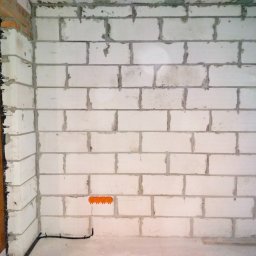 WJK Energia Wojciech Kubecki - Ściana z białych bloczków z widoczną puszką instalacyjną i kablem. Surowy stan, przygotowanie do prac wykończeniowych. Widoczne okno z drewnianą ramą.