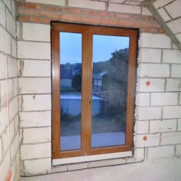 WJK Energia Wojciech Kubecki - Dwuskrzydłowe okno z brązową ramą, zamontowane w surowej ścianie z białych bloczków. Widoczne przewody elektryczne przy podłodze i fragment nieba za oknem.