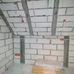 WJK Energia Wojciech Kubecki - Surowy stan poddasza z instalacją elektryczną w białych bloczkach, widoczne kable i puszki. Fragment izolacji na podłodze. Ujęcie z dołu, podkreślające surowość wnętrza.