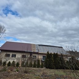 Dekarstwo - Częściowo pokryty dach budynku z brązową blachą i membraną dachową. Widoczne rusztowania i drzewa bez liści na pierwszym planie. Pochmurne niebo w tle.