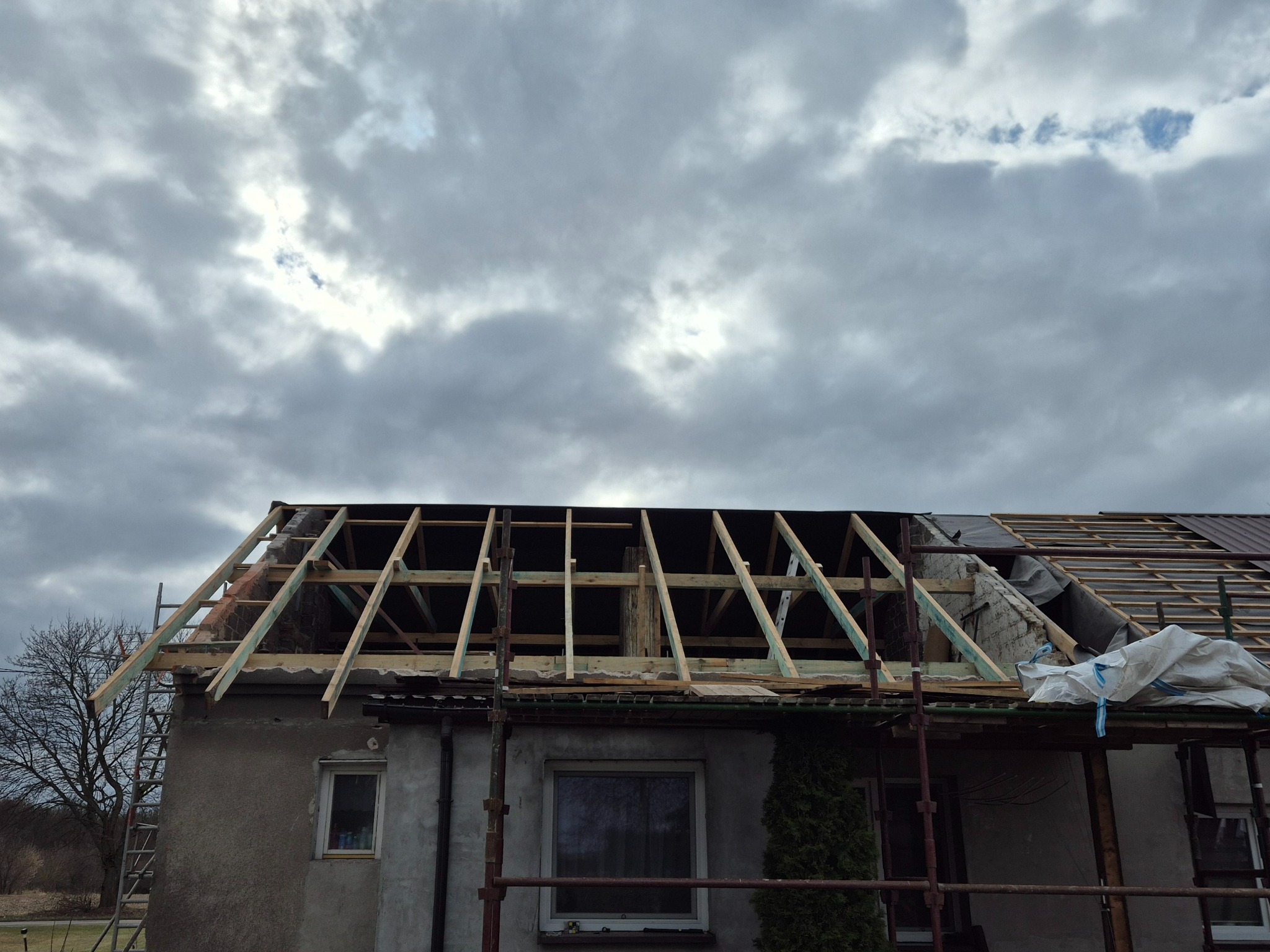 W trakcie budowy dachu domu jednorodzinnego. Widoczna drewniana konstrukcja więźby dachowej na tle pochmurnego nieba. Częściowo położone deski i folia dachowa.