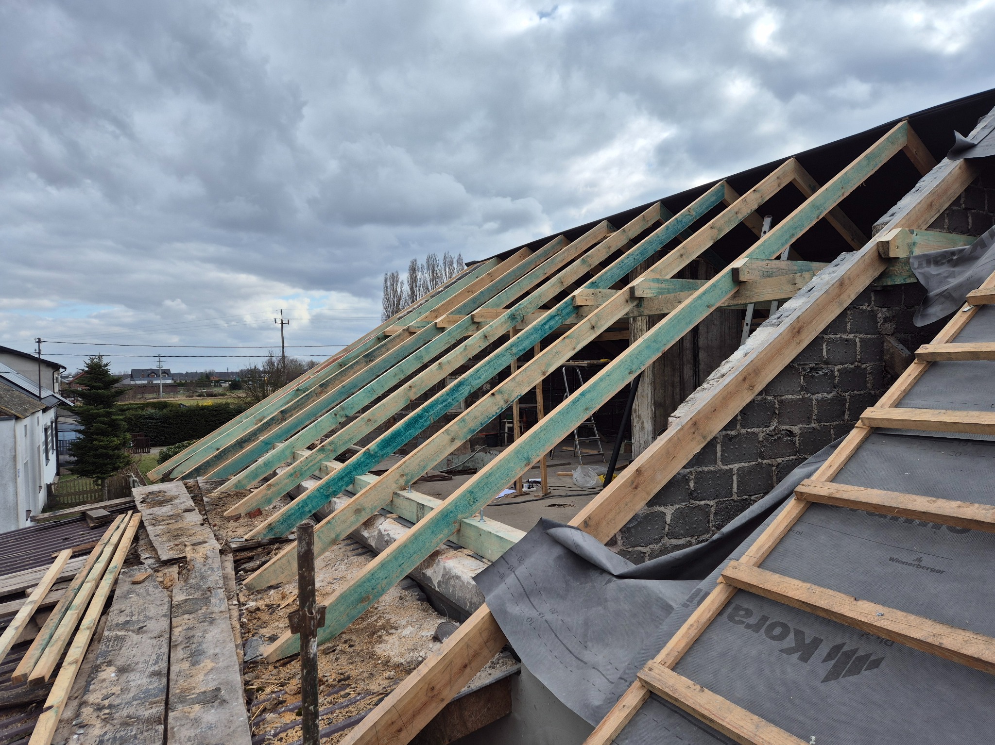 W trakcie budowy dachu: widoczna więźba dachowa zaimpregnowana na zielono, czarna membrana dachowa Kora i fragment starego dachu. W tle pochmurne niebo.