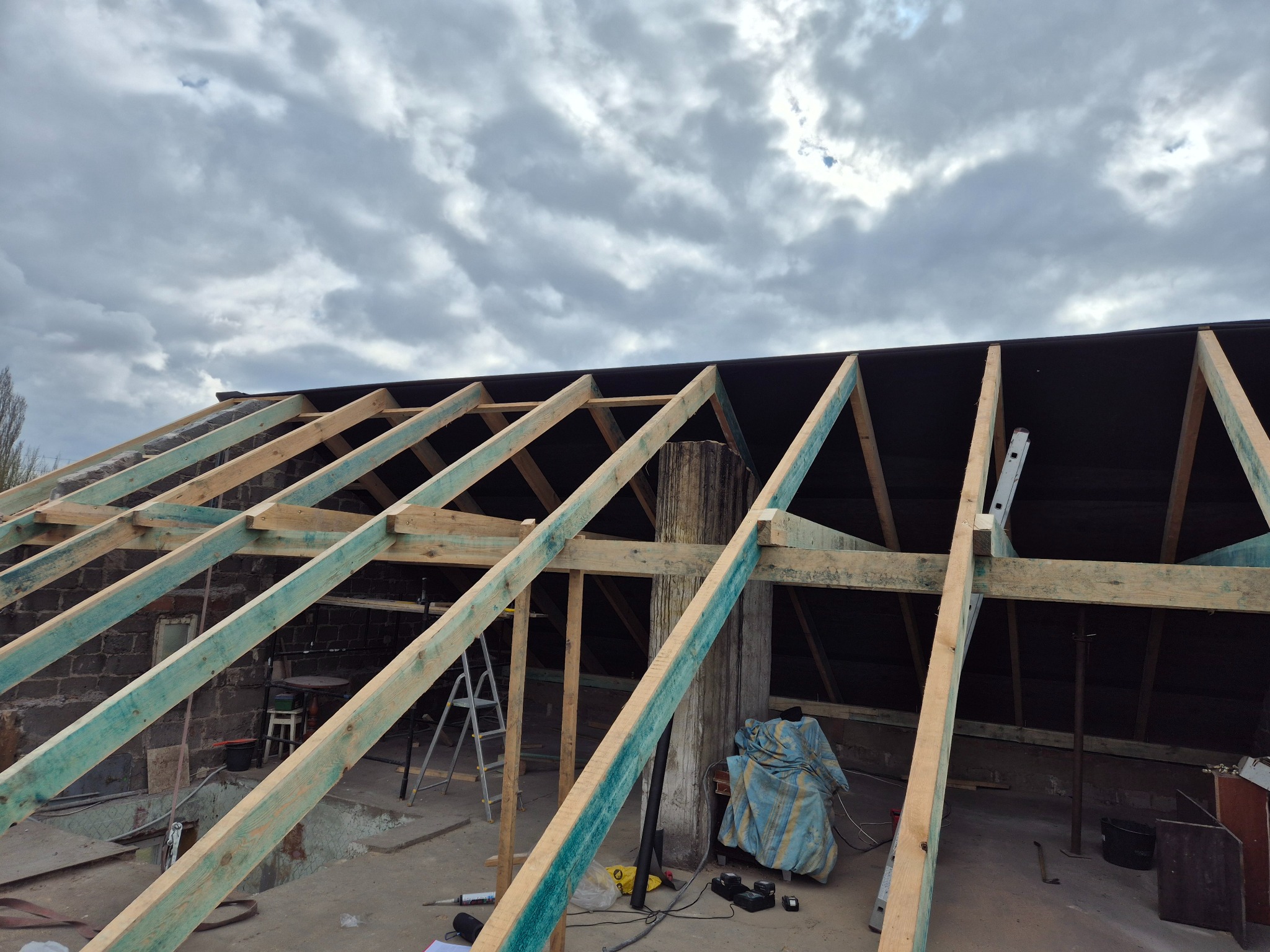 W trakcie budowy dachu: drewniana konstrukcja więźby dachowej z widoczną membraną, drabina i narzędzia na tle pochmurnego nieba. Ujęcie z dołu.