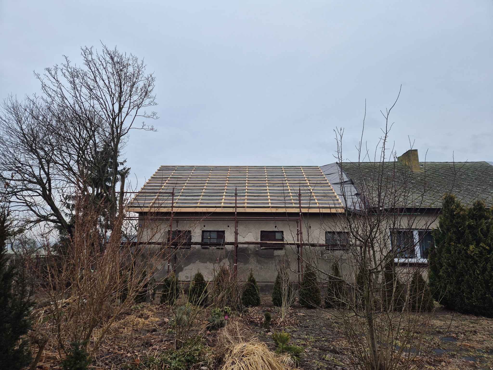 Budynek w trakcie prac dekarskich: widoczna konstrukcja dachu z łatami, częściowo pokryta membraną. Scaffolding przy ścianie. Drzewa bezlistne na pierwszym planie.