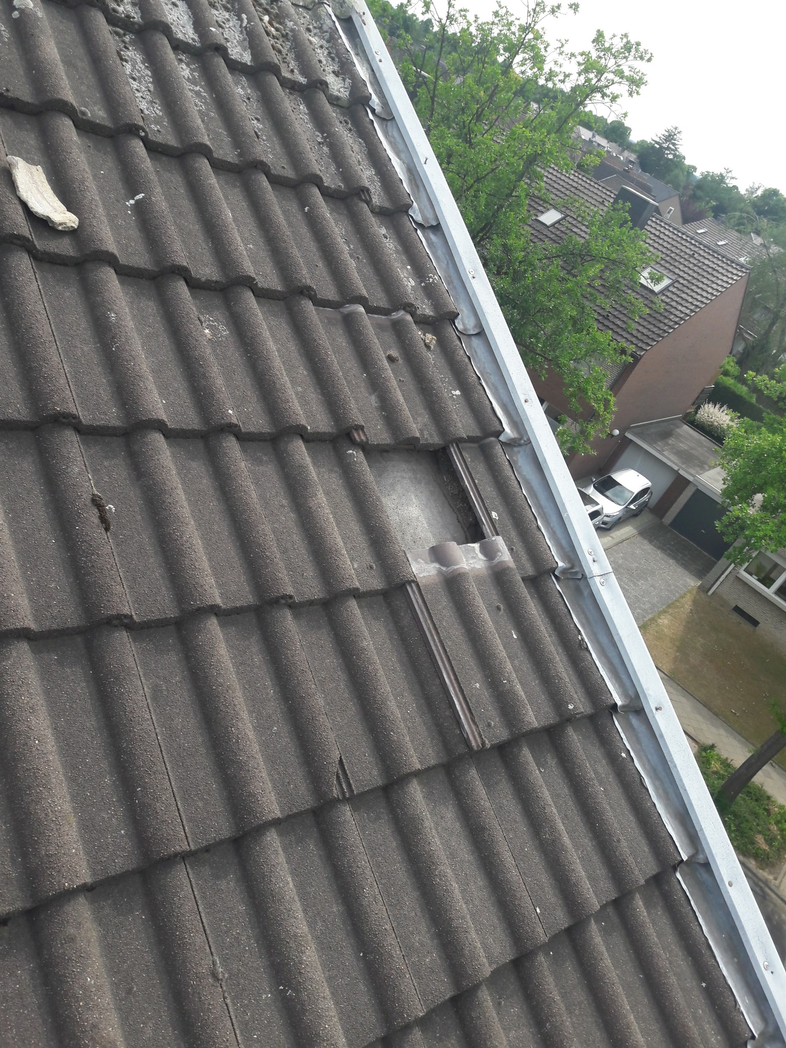 Uszkodzony dach z szarej dachówki ceramicznej, widoczny ubytek i fragment rynny. W tle zabudowa jednorodzinna i zieleń.