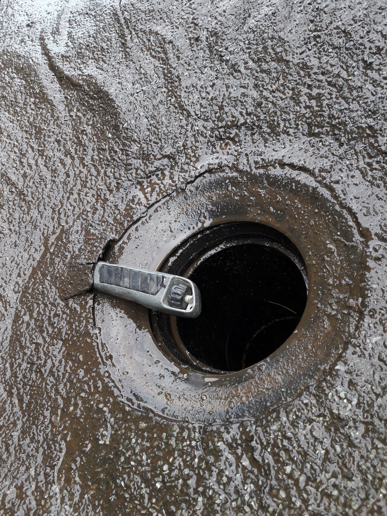 Otwarta studzienka kanalizacyjna na mokrej, betonowej powierzchni z narzędziem do podważania pokrywy. Widoczne wnętrze studzienki.