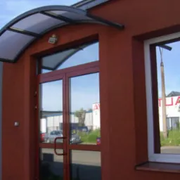 Wejście do budynku z brązowymi drzwiami i oknem, nad drzwiami daszek z przezroczystego tworzywa, widoczne odbicia w szybach.