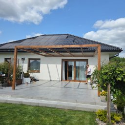 Timberview - Drewniana pergola przy domu z panelami słonecznymi, widok z trawnika. Mężczyzna wchodzi do domu przez przesuwne drzwi. Jasne niebo z chmurami.