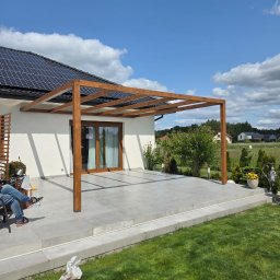 Timberview - Nowoczesna, drewniana pergola z belek na tarasie z szarych płyt, z widokiem na ogród i dom z panelami słonecznymi w słoneczny dzień. Częściowo widoczna osoba siedząca na krześle.