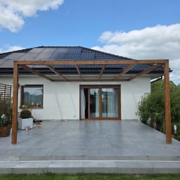 Timberview - Nowoczesna, brązowa pergola z drewna na szarym tarasie z płyt, przylegająca do białego domu z panelami słonecznymi na dachu. Błękitne niebo z chmurami w tle.
