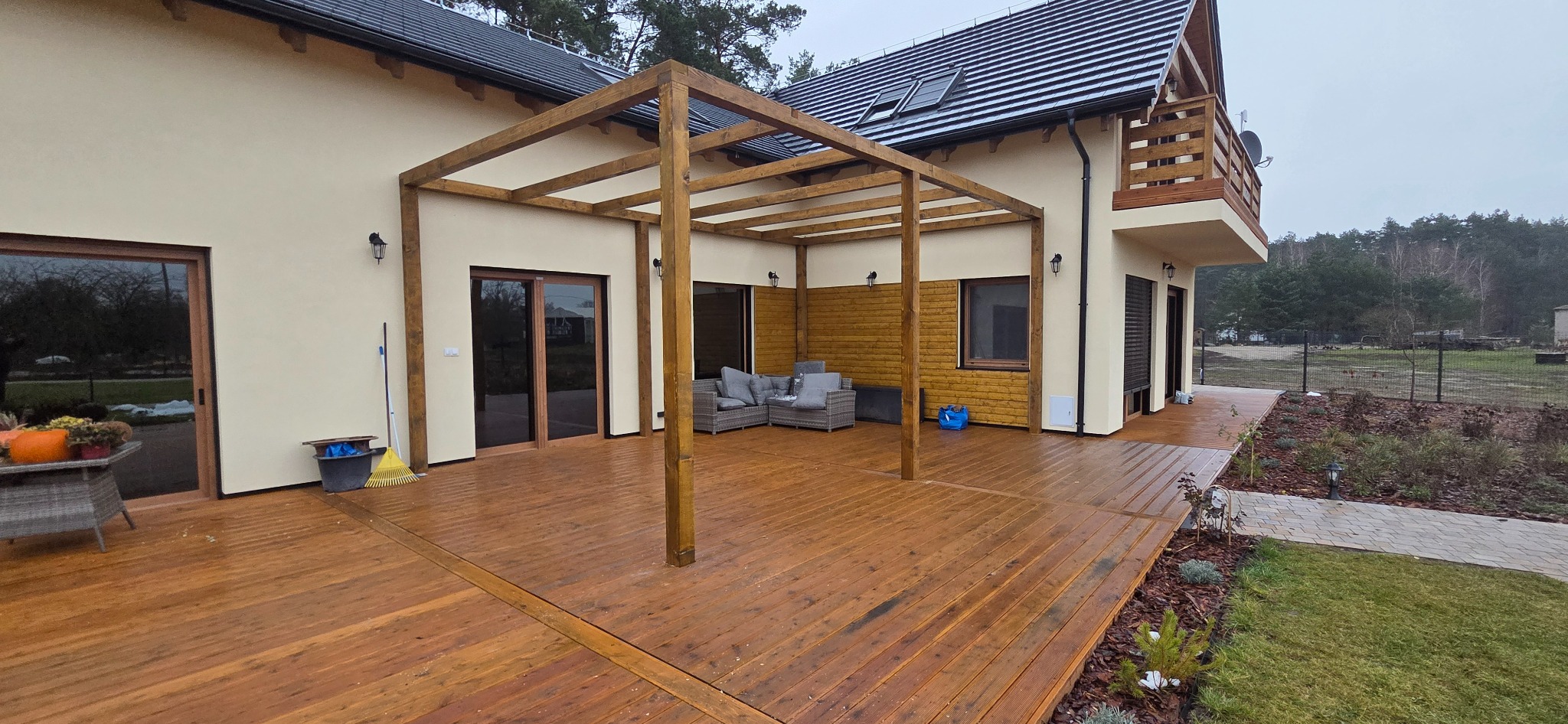 Przestronny taras z drewna z beżową elewacją domu i drewnianą pergolą. Na tarasie szara sofa ogrodowa. Widok na ogród i las w tle. Całość w naturalnej kolorystyce.