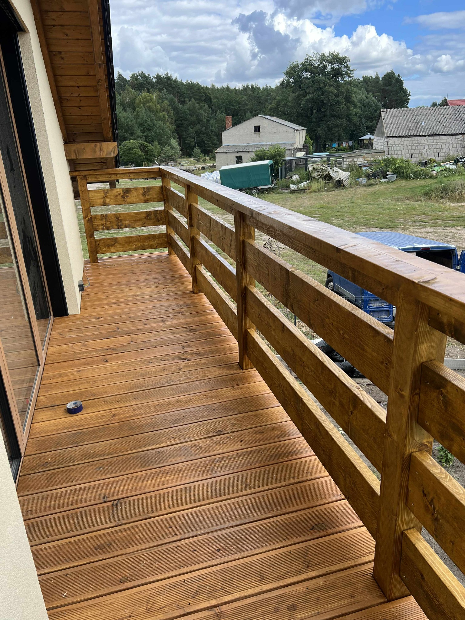 Balkon z drewnianą podłogą i balustradą z bejcowanego drewna, widok na zewnątrz. Na podłodze leży niebieska taśma malarska. W tle zabudowania gospodarcze i las.