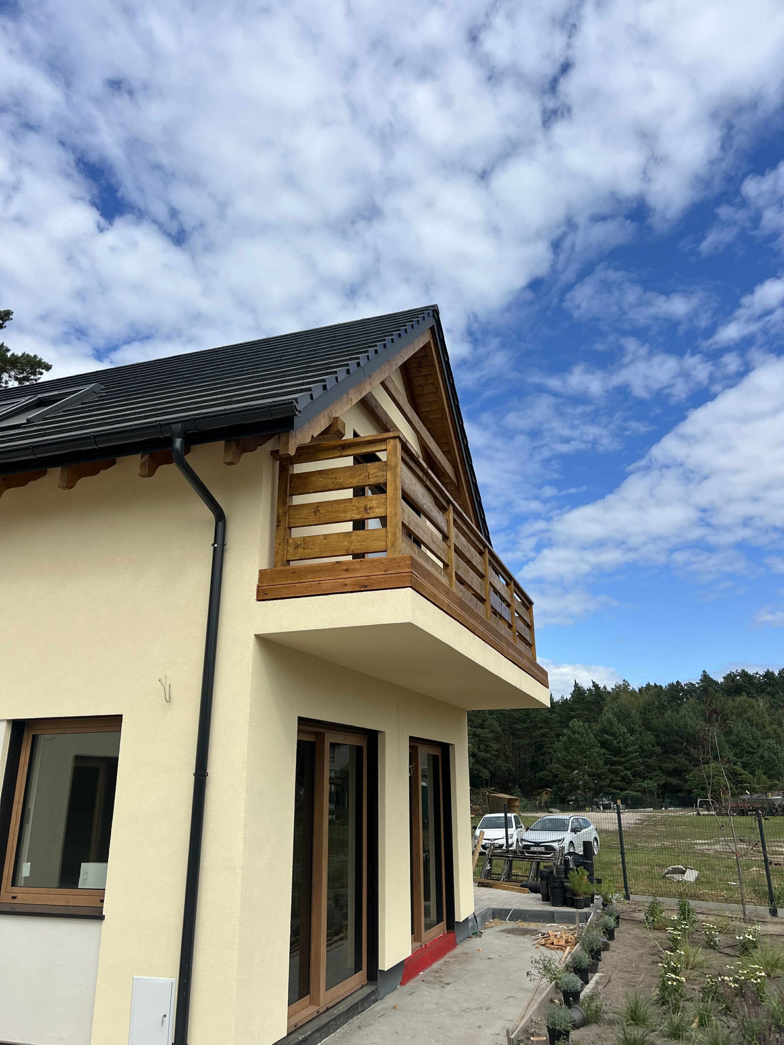 Nowoczesny dom z drewnianą balustradą balkonu, widok z dołu, błękitne niebo z chmurami w tle. Jasna elewacja, ciemna dachówka. Widoczne elementy wykończenia.