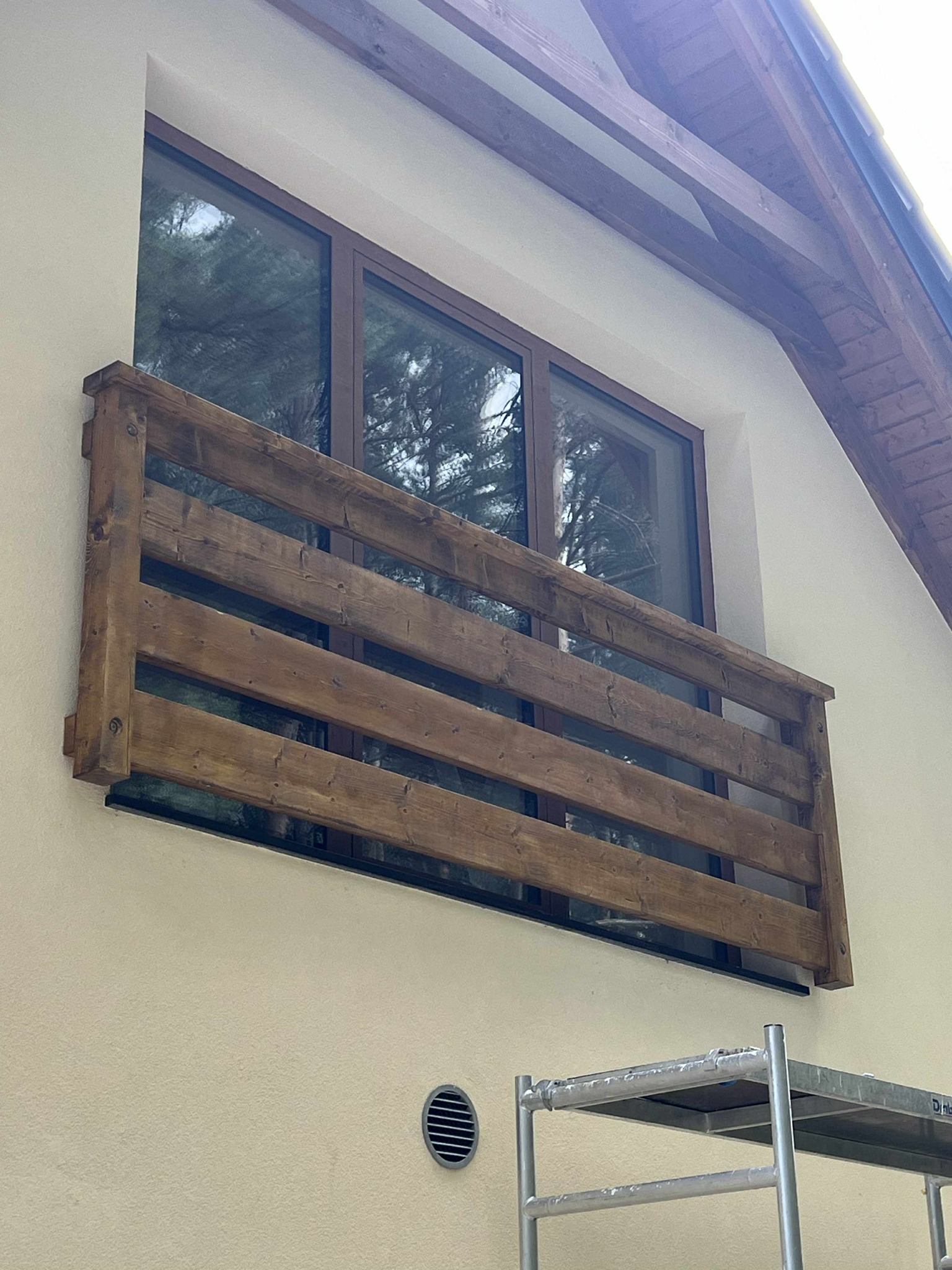 Drewniana balustrada balkonowa z bejcowanego drewna, zamontowana na elewacji budynku z oknem. Widoczne rusztowanie w dolnej części kadru.