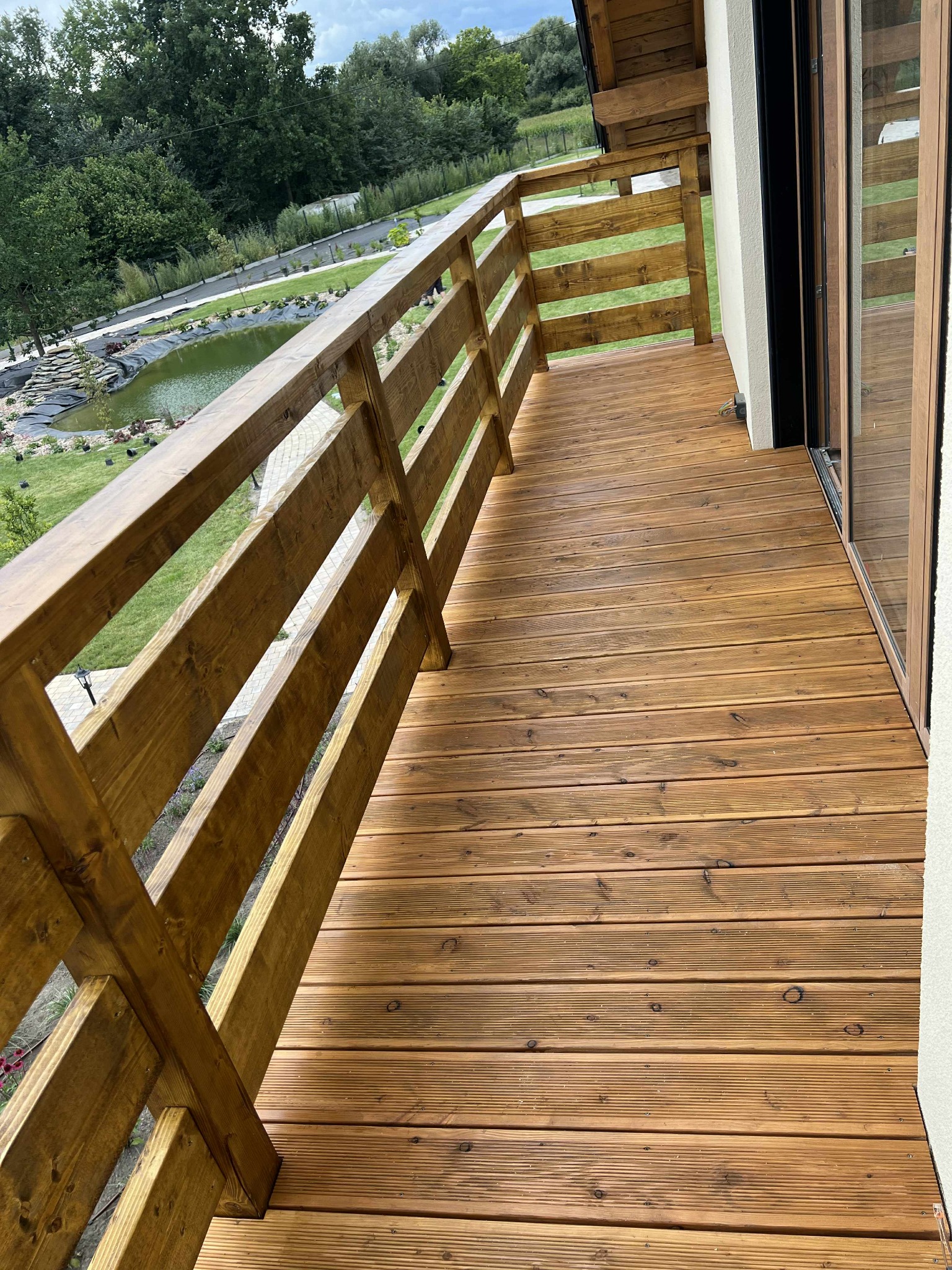 Balkon z drewnianą podłogą i balustradą, widok na ogród z oczkiem wodnym. Naturalne oświetlenie, ciepłe odcienie drewna. Detal wykończenia balkonu.