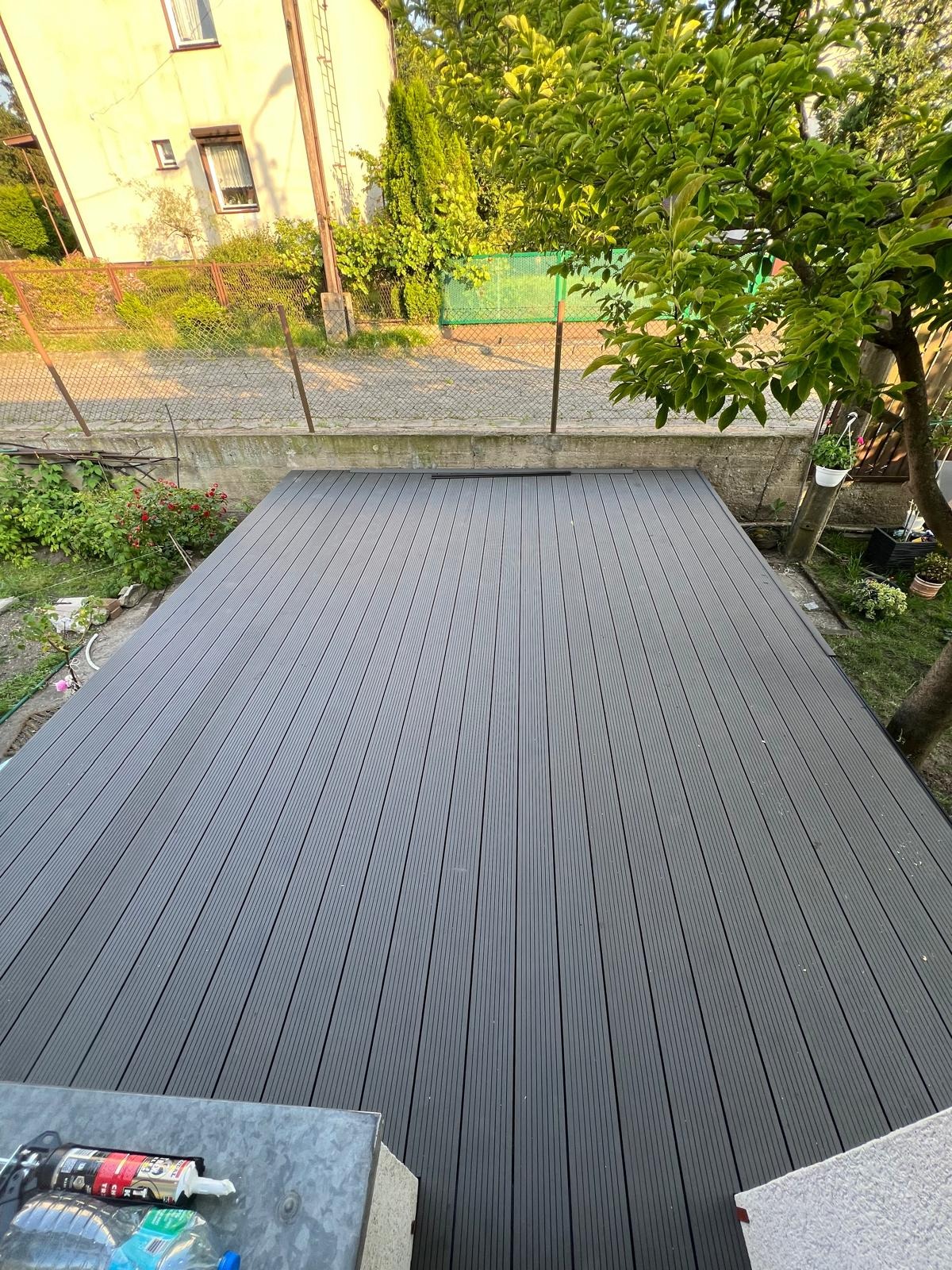 Szary taras kompozytowy z widokiem z góry, otoczony zielenią ogrodu i elementami architektury. Deski ułożone równolegle, tworząc uporządkowaną powierzchnię.