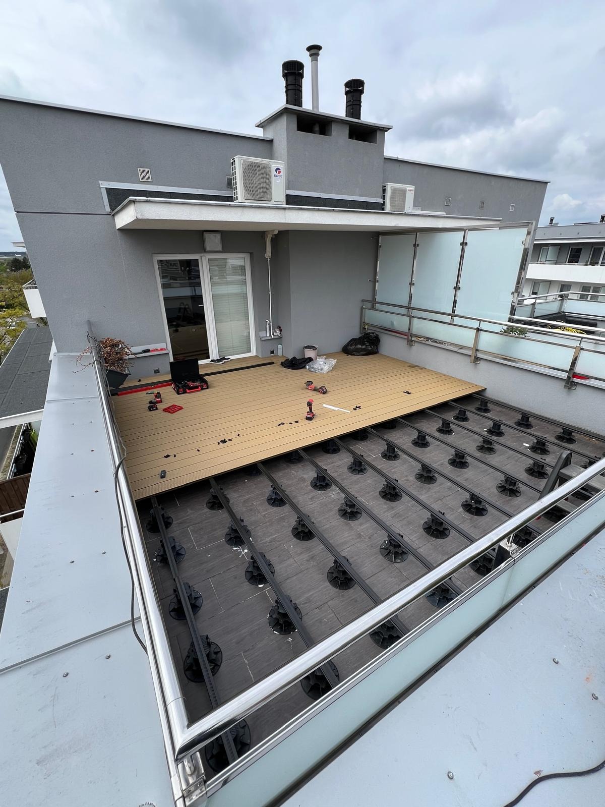 Montaż tarasu kompozytowego na balkonie z widoczną konstrukcją wsporczą i częściowo ułożonymi deskami. Widoczne narzędzia i akcesoria montażowe. Balkon z szarą elewacją.