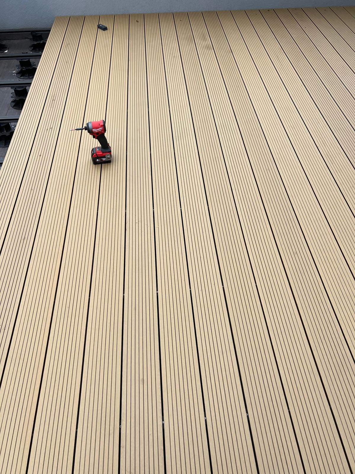 Jasny taras kompozytowy z widocznym wkrętakiem akumulatorowym, ukazujący etap montażu desek. Widoczne elementy mocujące i regularny układ desek.