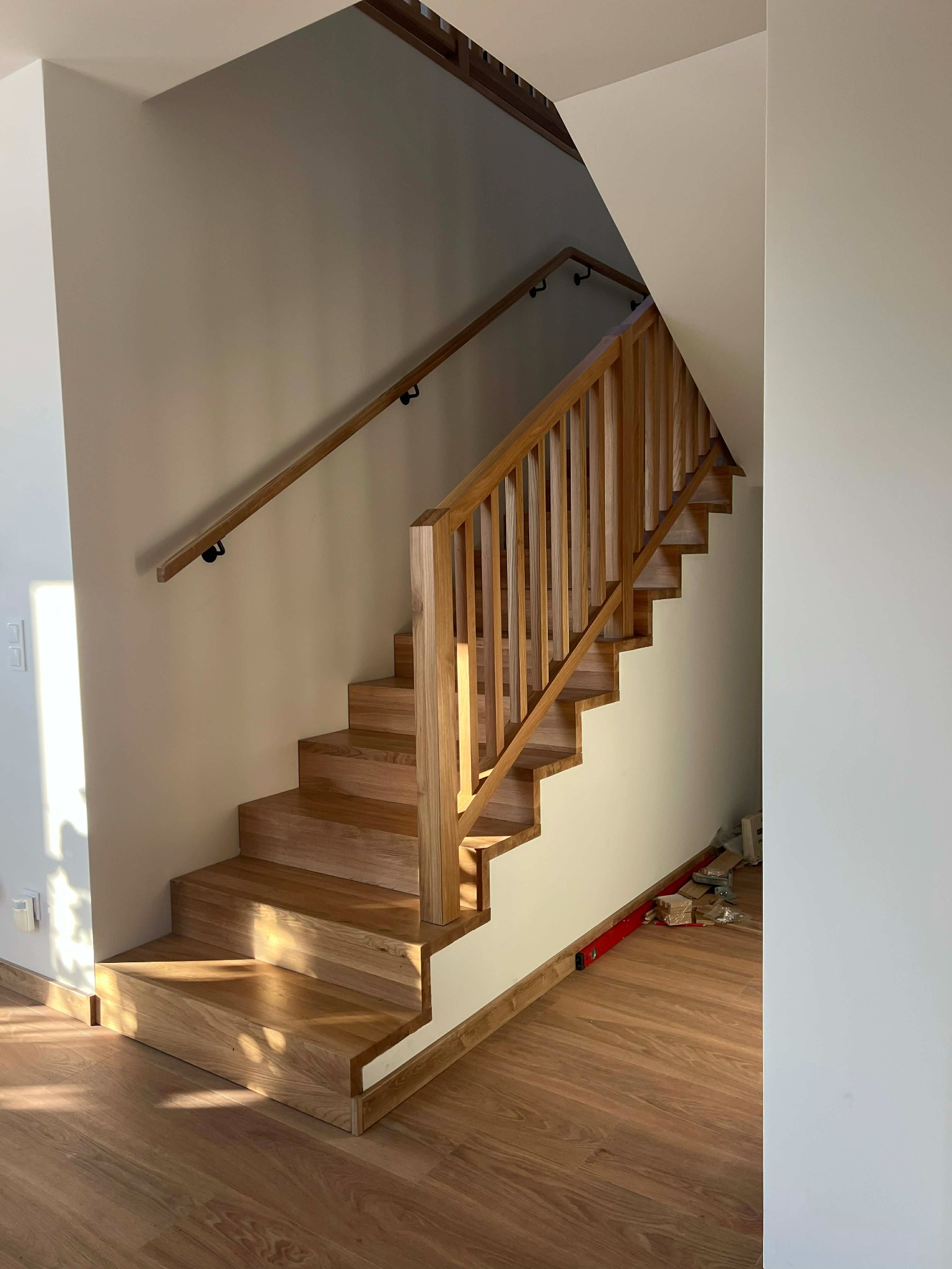 Jasne, drewniane schody z balustradą, prowadzące na górę. Widoczne detale wykończenia i naturalne oświetlenie. Częściowo widoczne narzędzia na podłodze.