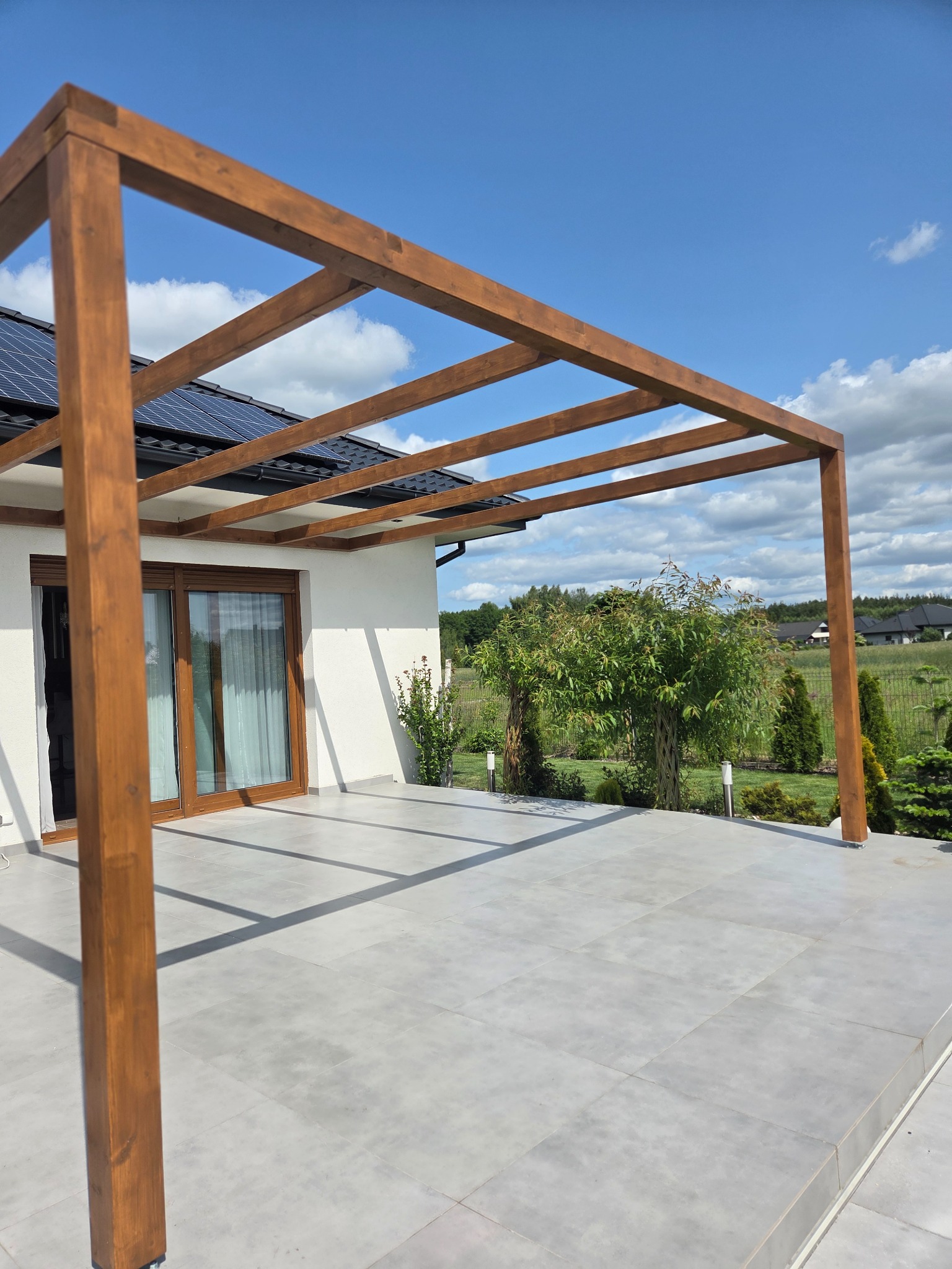 Nowoczesna pergola z drewna na tarasie z szarych płyt, z widokiem na ogród i dom z panelami słonecznymi. Jasne, słoneczne oświetlenie.
