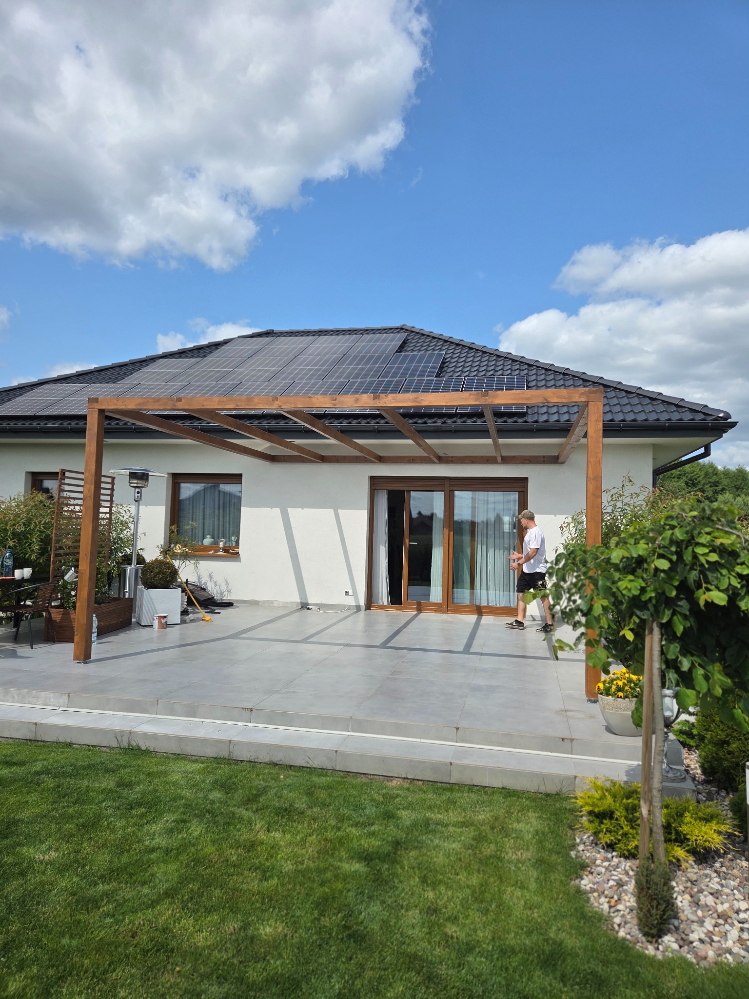 Drewniana pergola przy domu z panelami słonecznymi, widok z trawnika. Mężczyzna wchodzi do domu przez przesuwne drzwi. Jasne niebo z chmurami.