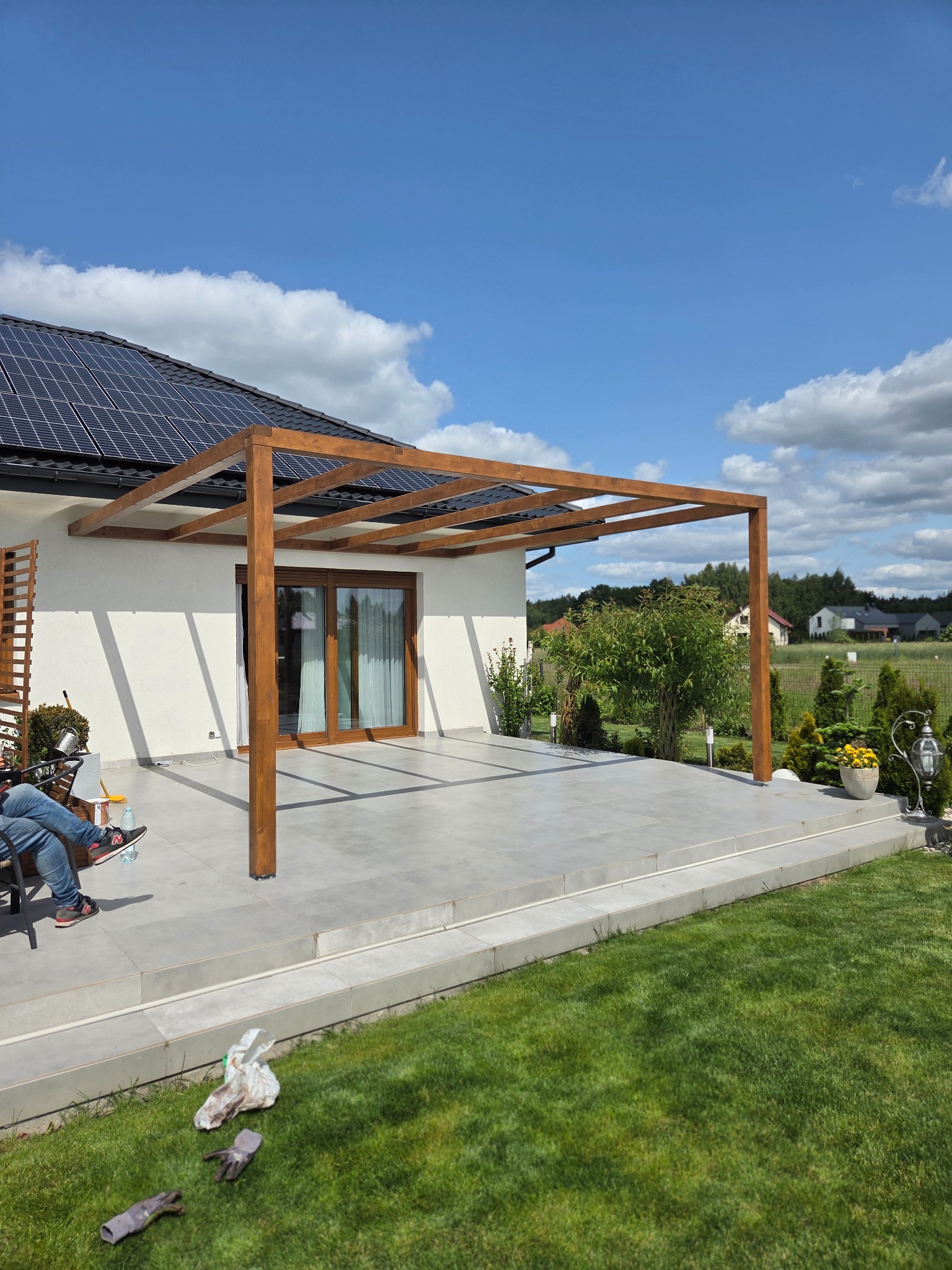 Nowoczesna, drewniana pergola z belek na tarasie z szarych płyt, z widokiem na ogród i dom z panelami słonecznymi w słoneczny dzień. Częściowo widoczna osoba siedząca na krześle.