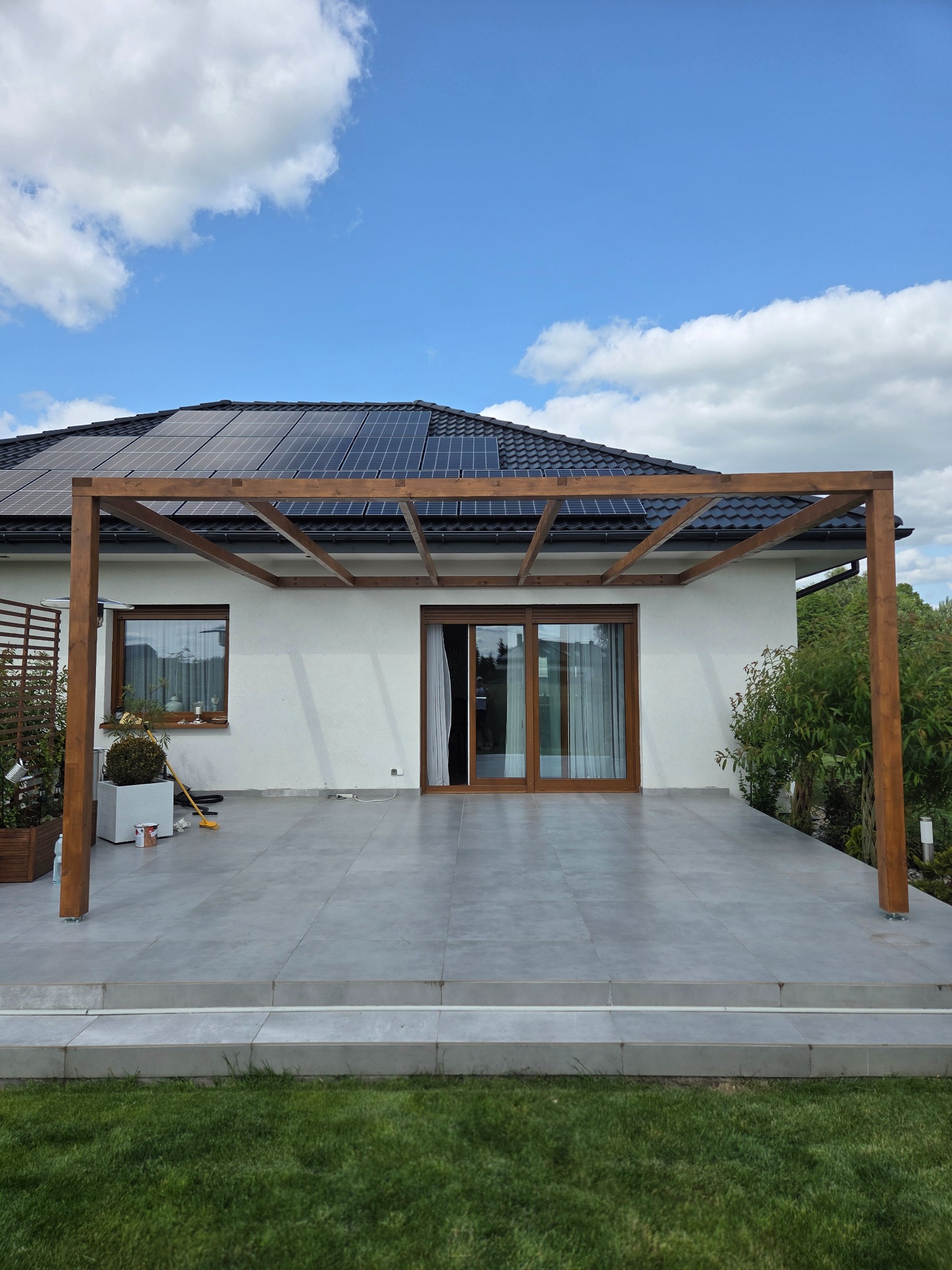 Nowoczesna, brązowa pergola z drewna na szarym tarasie z płyt, przylegająca do białego domu z panelami słonecznymi na dachu. Błękitne niebo z chmurami w tle.