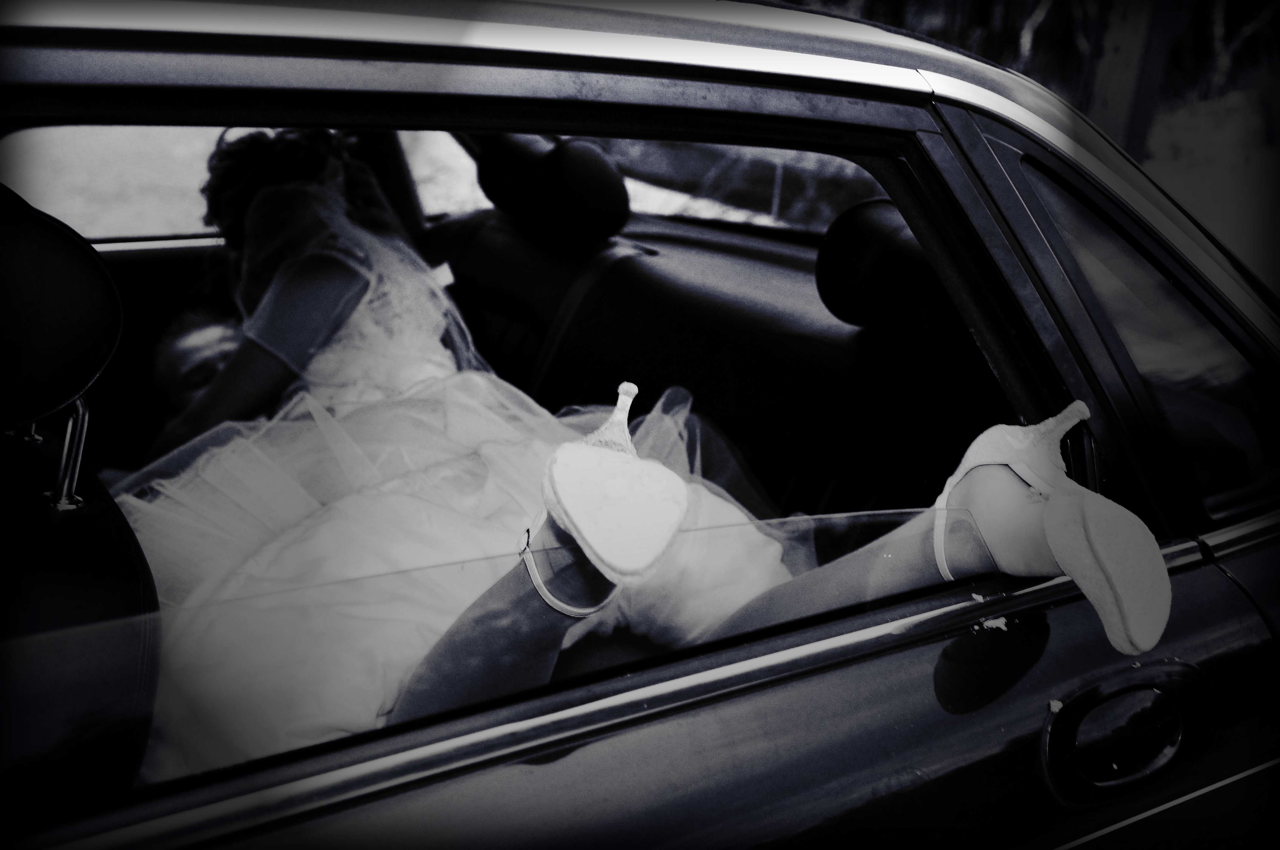 Panna młoda w sukni ślubnej i białych butach, siedząca w samochodzie, widoczna przez otwarte okno; czarno-białe zdjęcie.