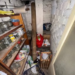 Jakub Marczak - Zagracona piwnica z regałem pełnym słoików, workami i starymi przedmiotami. Białe ściany z cegły i betonowy sufit. Widoczny wózek na zakupy.