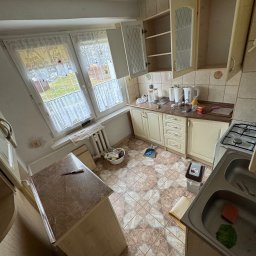 Jakub Marczak - Kuchnia przed remontem: widok z góry na starą zabudowę, płytki podłogowe w geometryczny wzór, okno z firankami i widoczne ślady użytkowania.