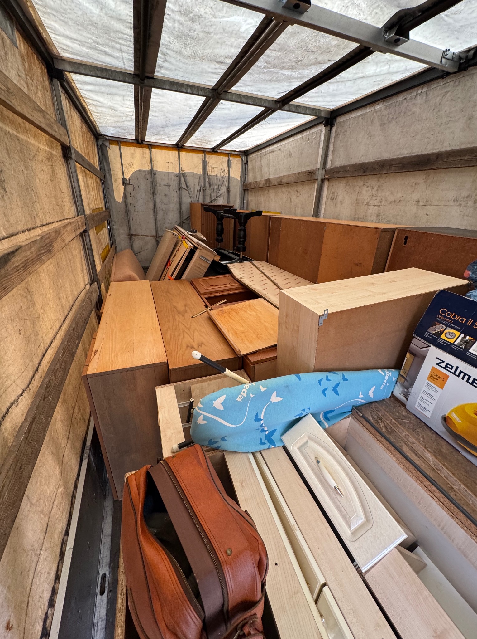 Wnętrze skrzyni ładunkowej wypełnionej meblami i sprzętami AGD, gotowymi do transportu. Widoczna torba podróżna i kartony z odkurzaczami.