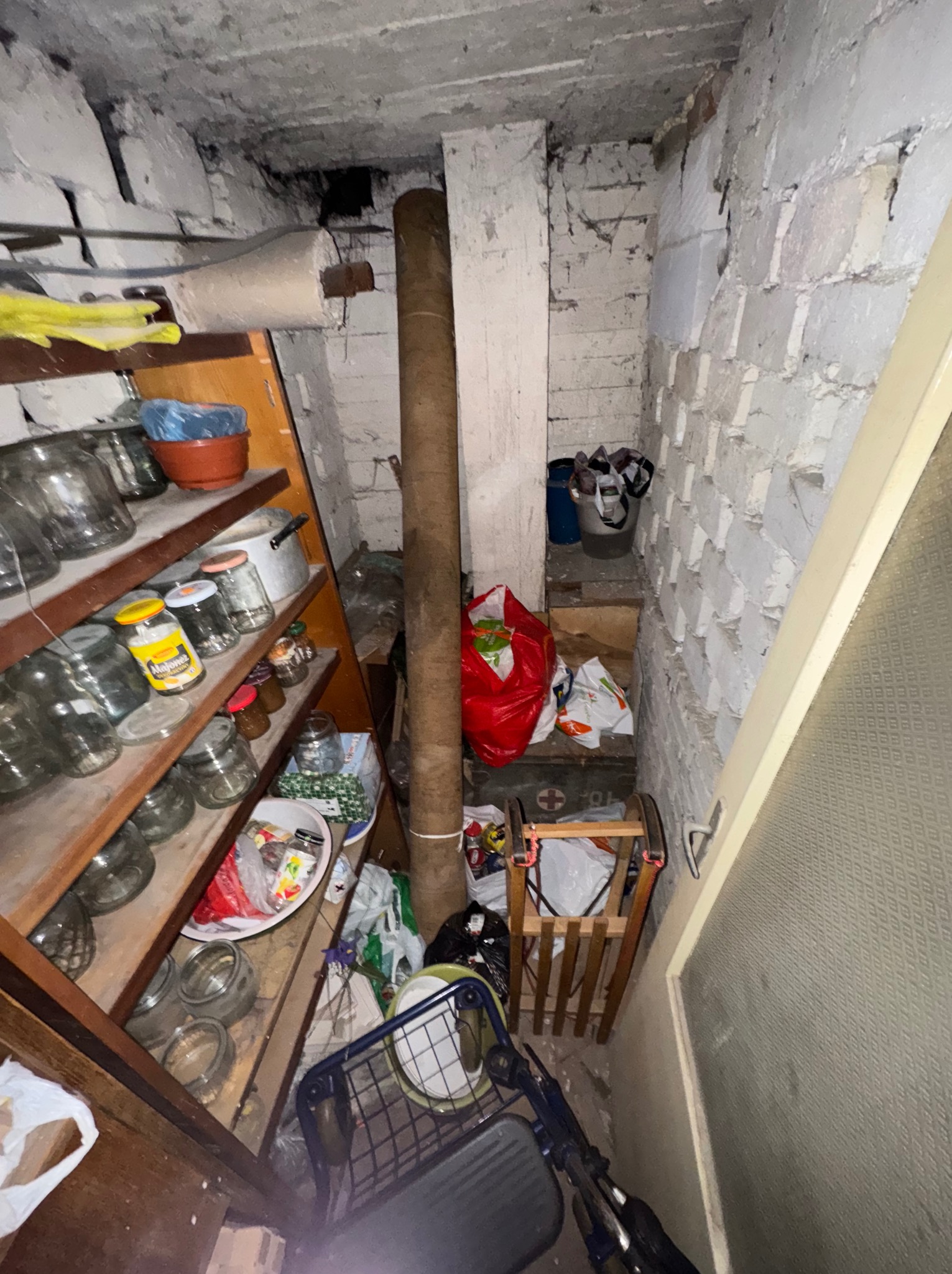 Zagracona piwnica z regałem pełnym słoików, workami i starymi przedmiotami. Białe ściany z cegły i betonowy sufit. Widoczny wózek na zakupy.