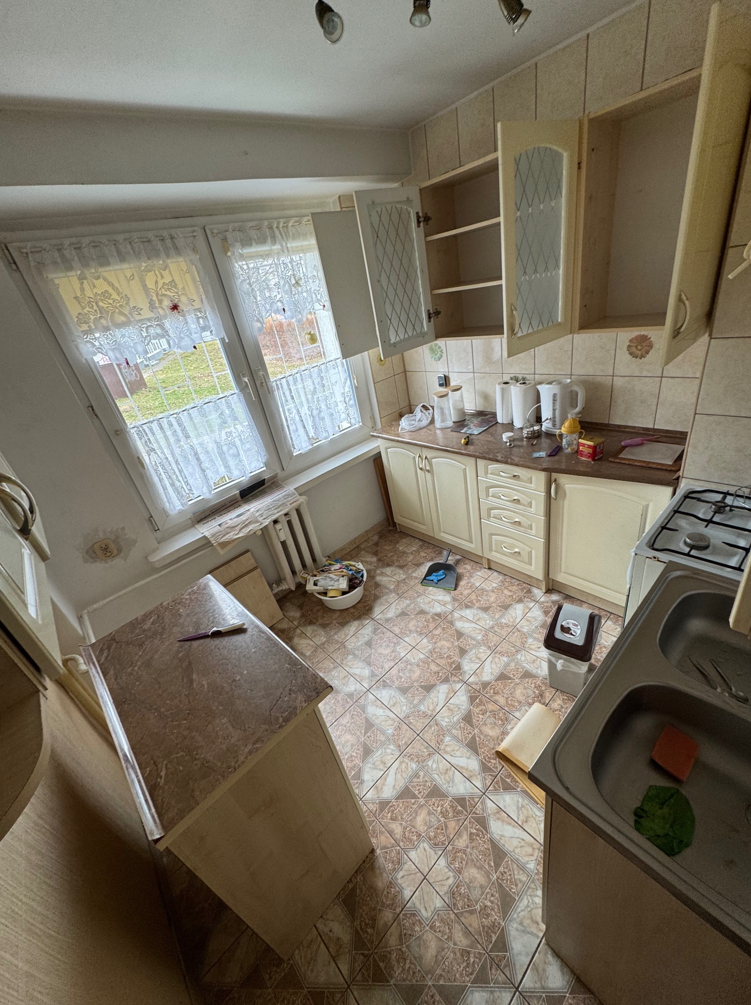 Kuchnia przed remontem: widok z góry na starą zabudowę, płytki podłogowe w geometryczny wzór, okno z firankami i widoczne ślady użytkowania.