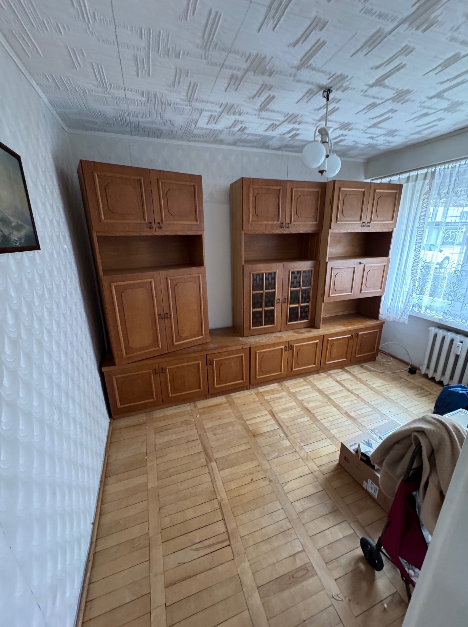 Pokój z drewnianą podłogą i meblościanką przed opróżnianiem. Widoczny wózek transportowy i karton, sugerujące przygotowania do wyniesienia wyposażenia.