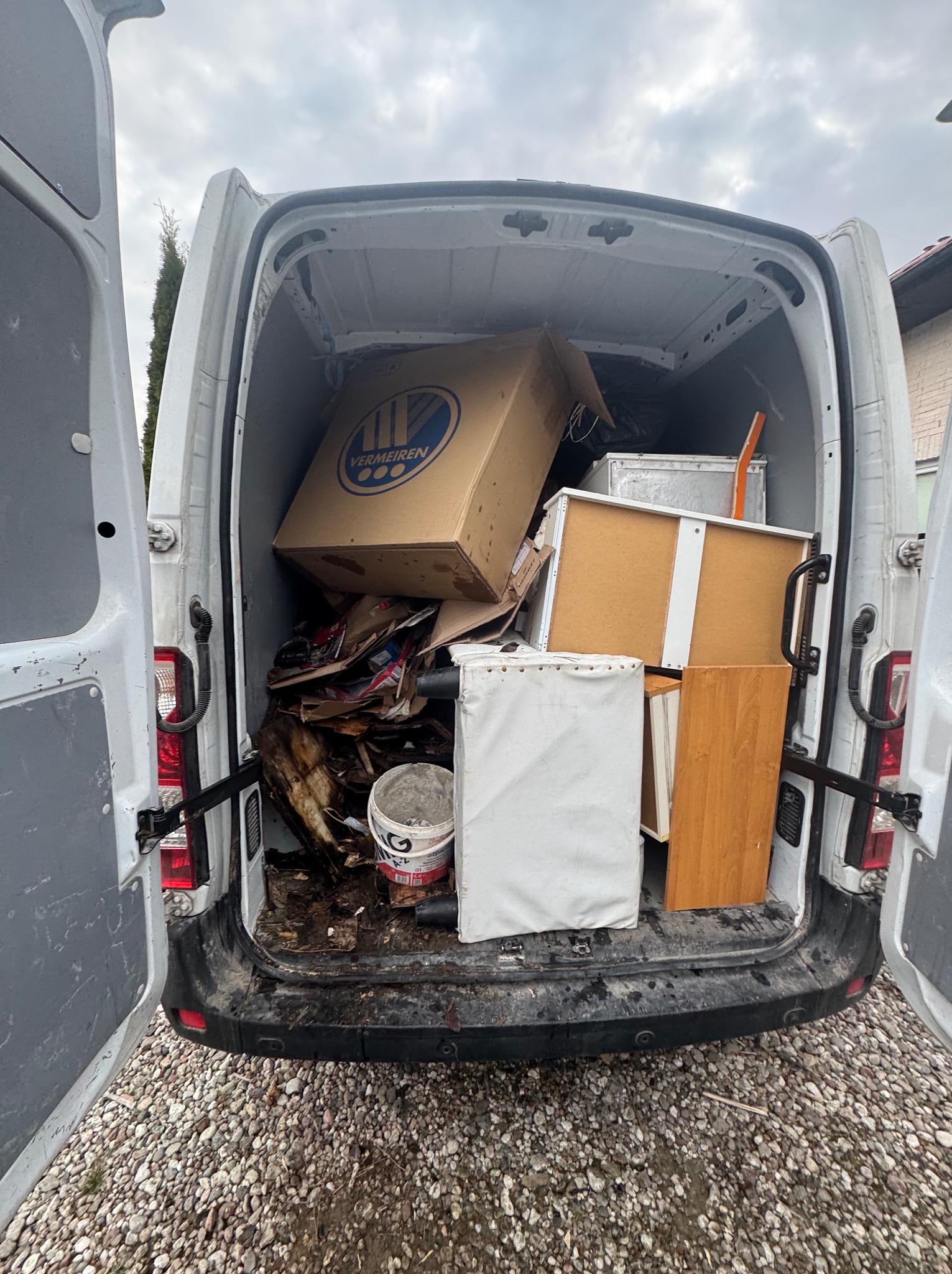 Załadunek białego busa kartonami, meblami i odpadami. Widoczne otwarte tylne drzwi, kamieniste podłoże i fragment budynku w tle. Częściowy widok zawartości.