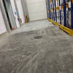 Mediclean Profesjonalne mycie ciśnieniowe - Betonowa posadzka w hali magazynowej po czyszczeniu, widoczne regały magazynowe i ślady po maszynach czyszczących. Szara posadzka, żółte barierki ochronne.