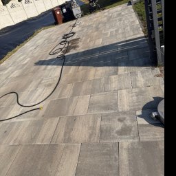 Mediclean Profesjonalne mycie ciśnieniowe - Kostka brukowa po czyszczeniu w Chrzanowie. Widoczny wąż ciśnieniowy i urządzenie czyszczące. Czysta, szara kostka na podjeździe, w tle ogrodzenie i zieleń.