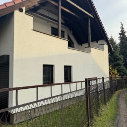 Mediclean Profesjonalne mycie ciśnieniowe - Dwupiętrowy dom z białymi ścianami i drewnianym balkonem, widziany zza metalowego ogrodzenia. Widoczne okna i fragment dachu. Tło stanowią drzewa.