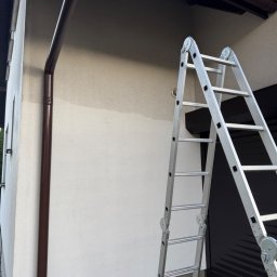 Mediclean Profesjonalne mycie ciśnieniowe - Fragment elewacji domu po częściowym umyciu, z widoczną różnicą w kolorze. Aluminiowa drabina oparta o ścianę, brązowa rynna wzdłuż krawędzi. Ujęcie z dołu.