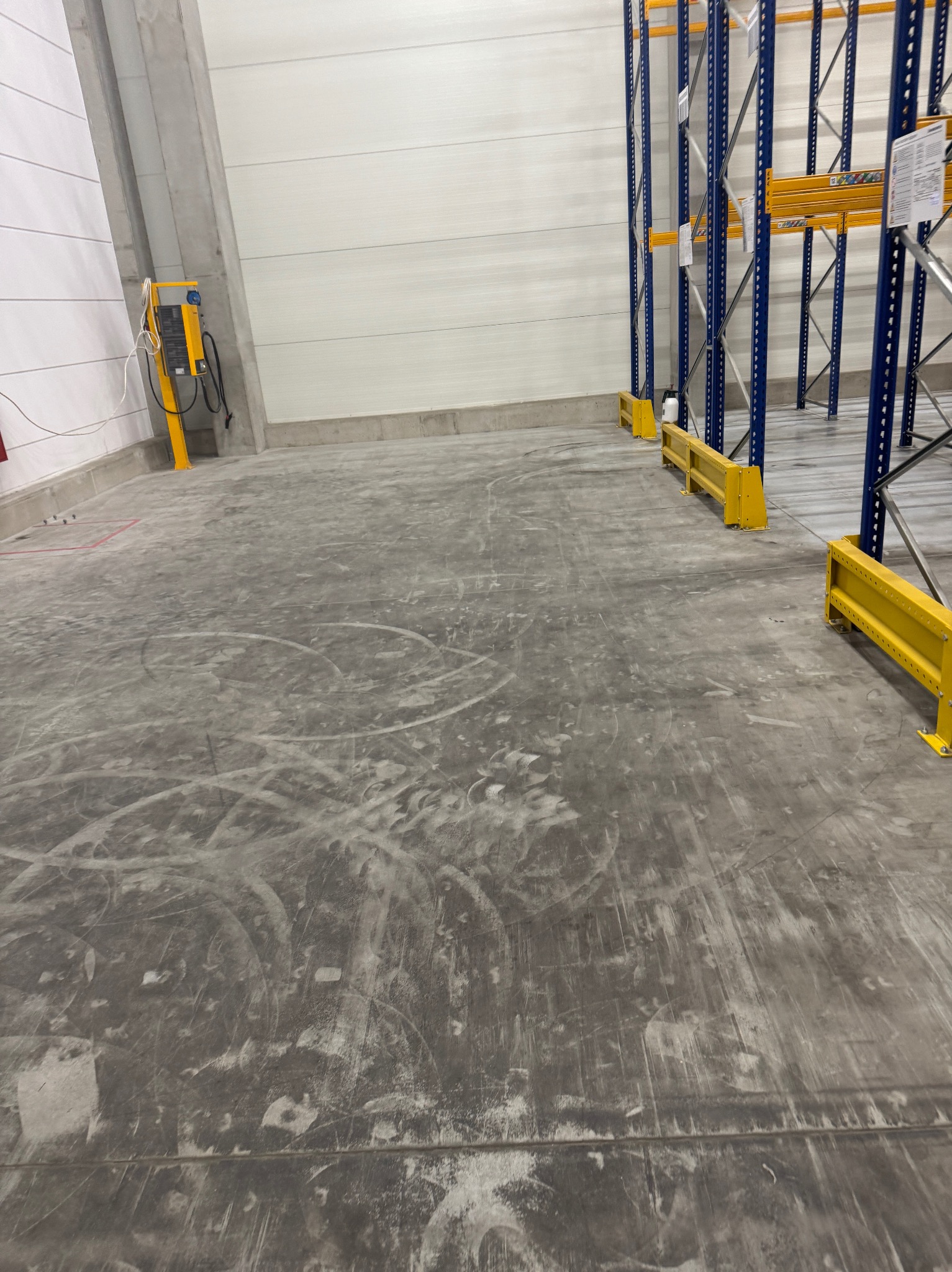 Betonowa posadzka w hali magazynowej przed sprzątaniem, widoczne ślady po wózkach widłowych i regałach. W tle biała ściana i fragment regałów magazynowych.