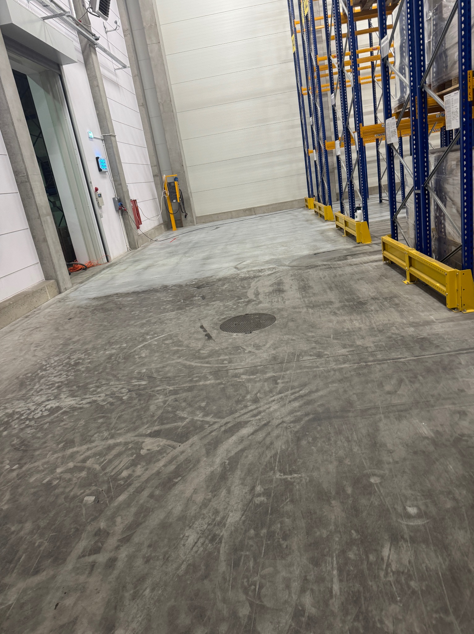 Betonowa posadzka w hali magazynowej po czyszczeniu, widoczne regały magazynowe i ślady po maszynach czyszczących. Szara posadzka, żółte barierki ochronne.