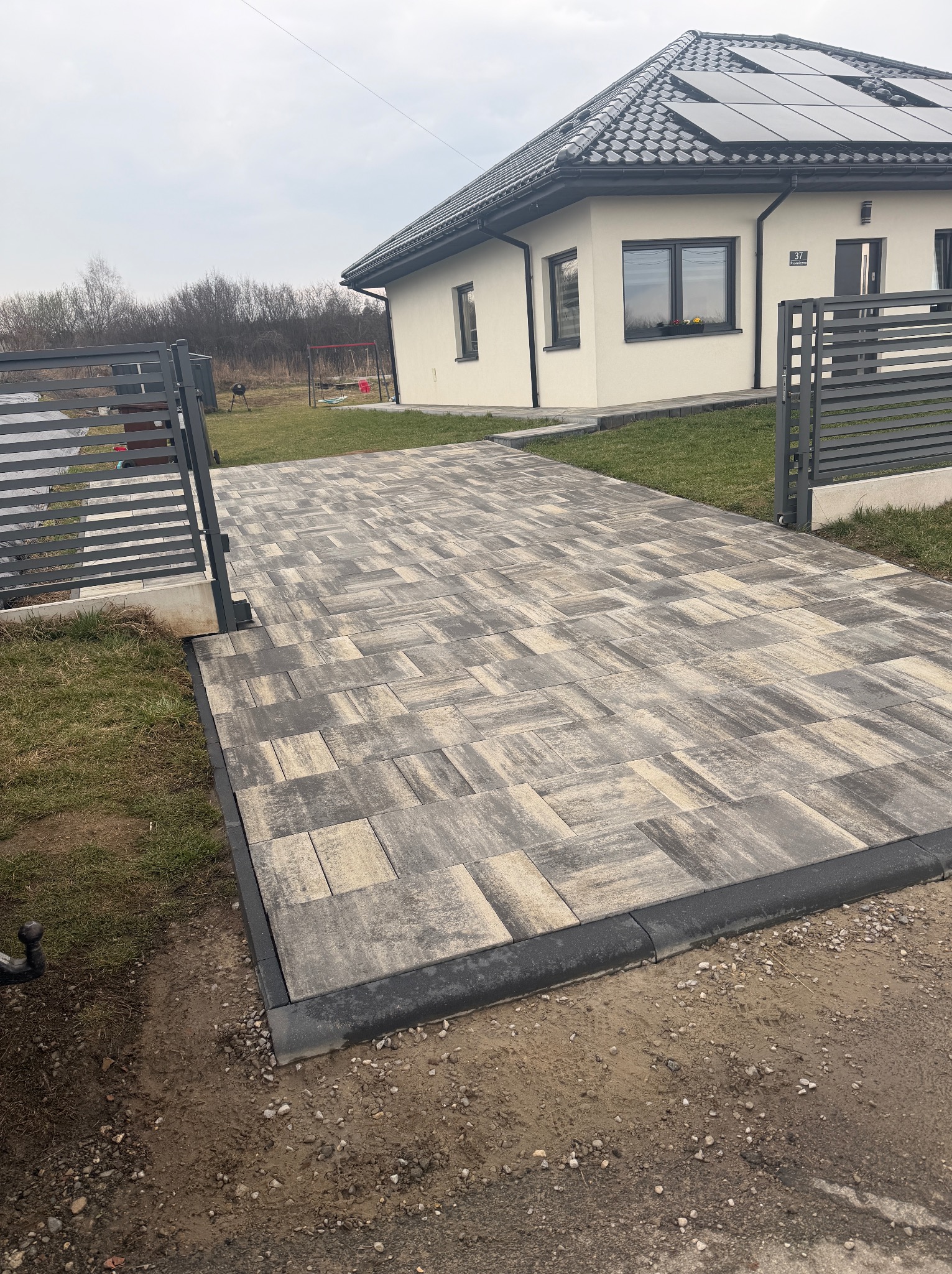 Ułożona kostka brukowa na podjeździe w Chrzanowie, widok z poziomu gruntu. Na dachu domu panele fotowoltaiczne. Otwarta brama wjazdowa.